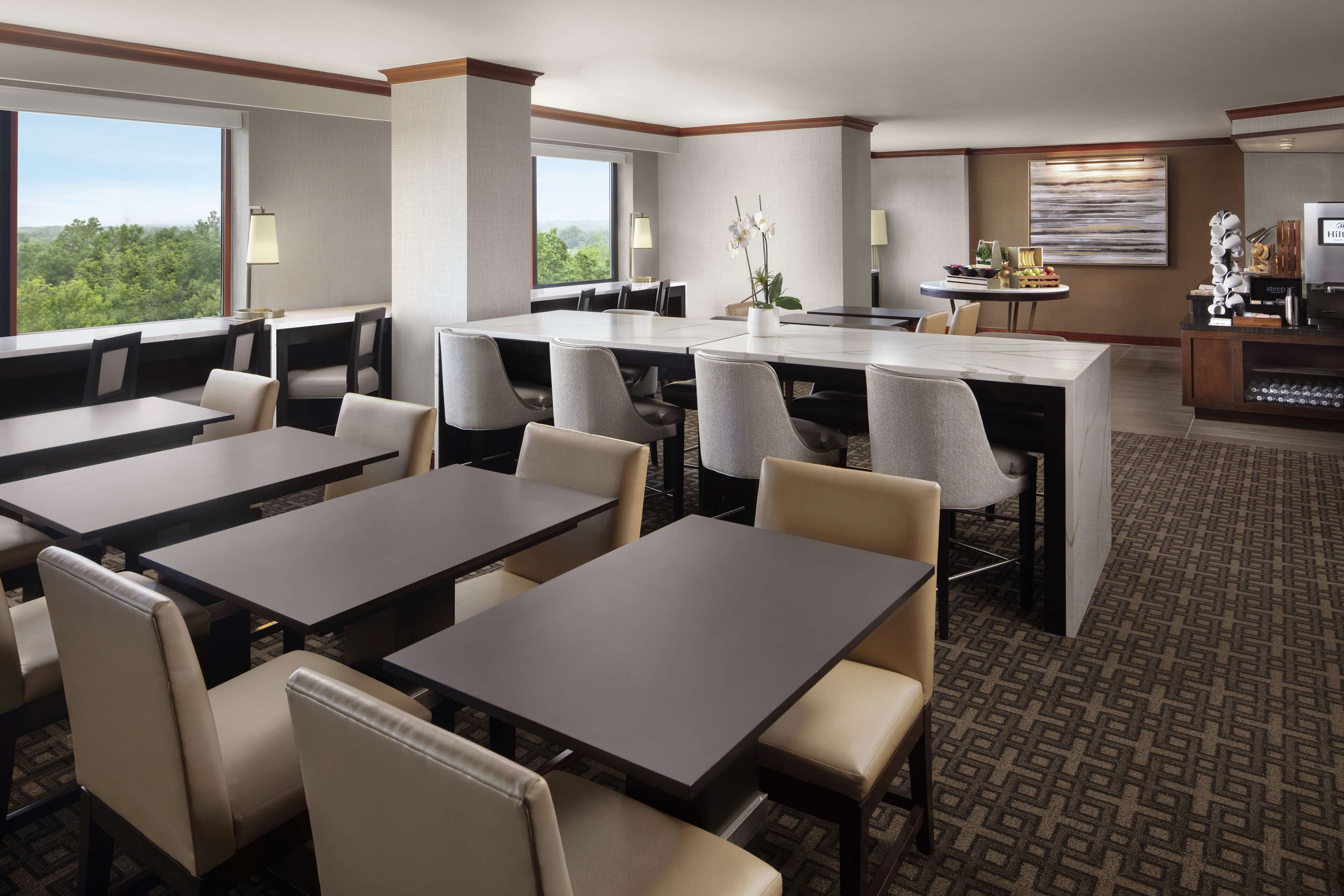 A hotel dining area with multiple tables and chairs, a central island with a vase of flowers, and a television on a stand.