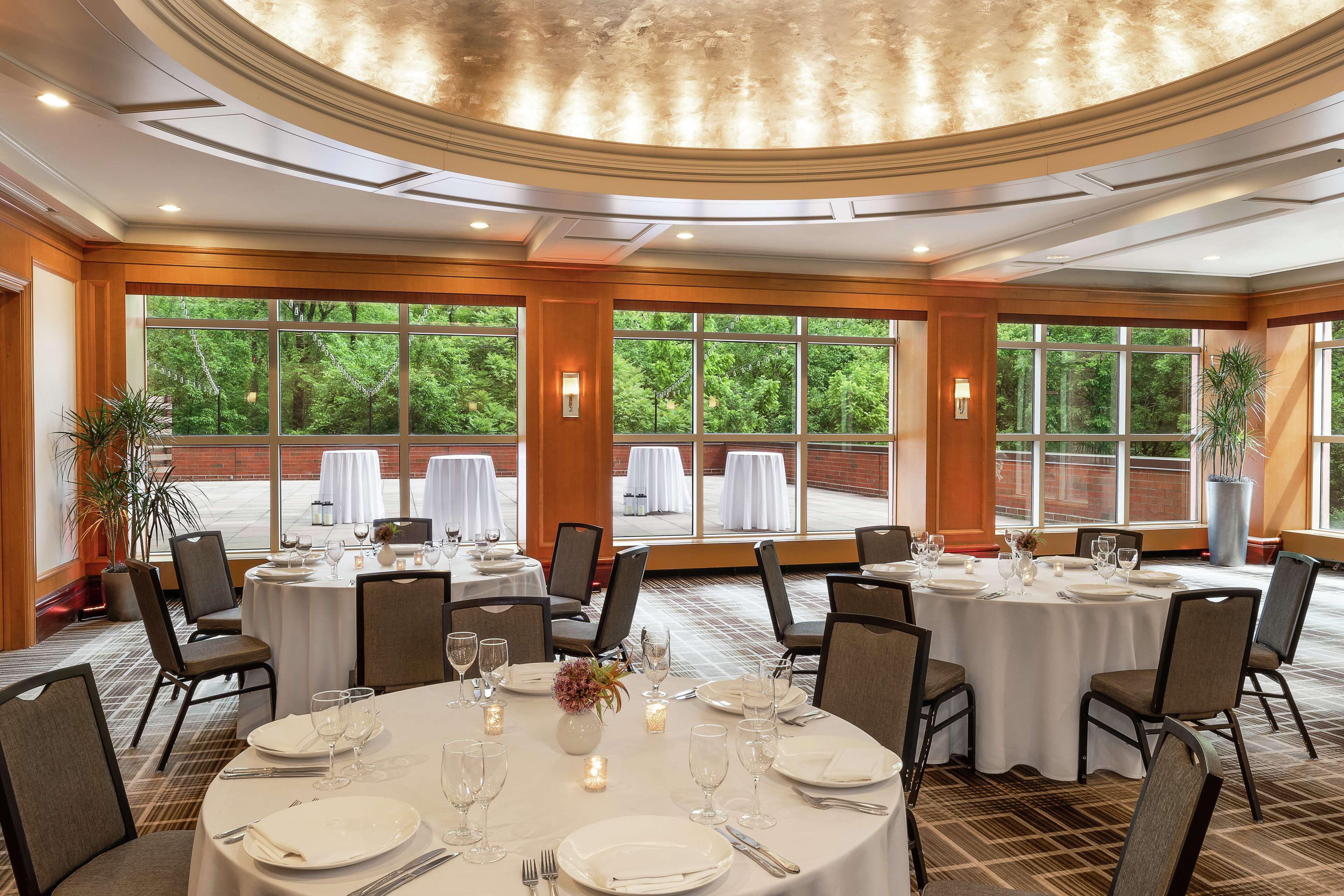 A banquet hall with round tables set for dining, large windows with a view of greenery, and a polished ceiling.