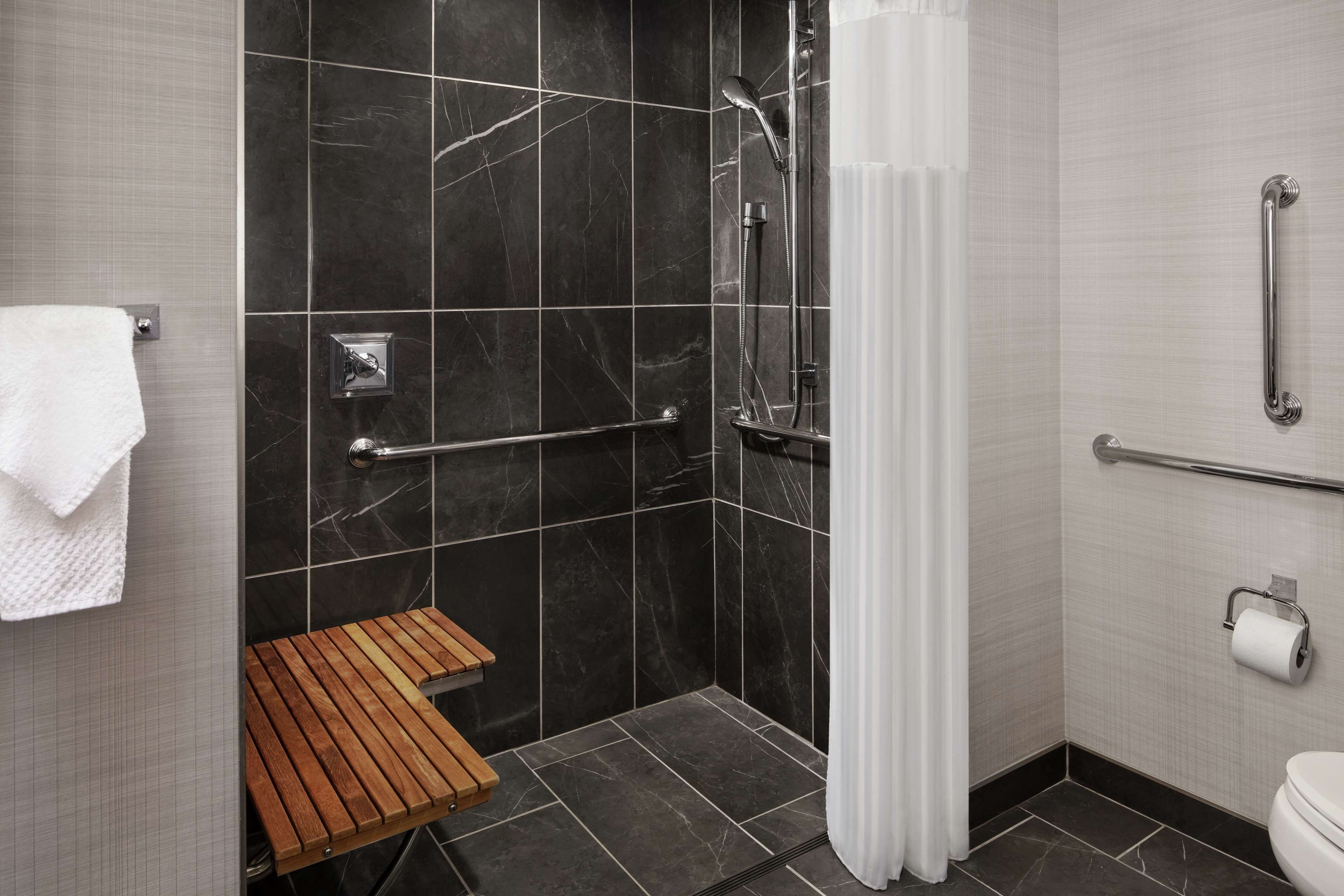 A modern bathroom with a shower, grab bars, a wooden bench, and a toilet.
