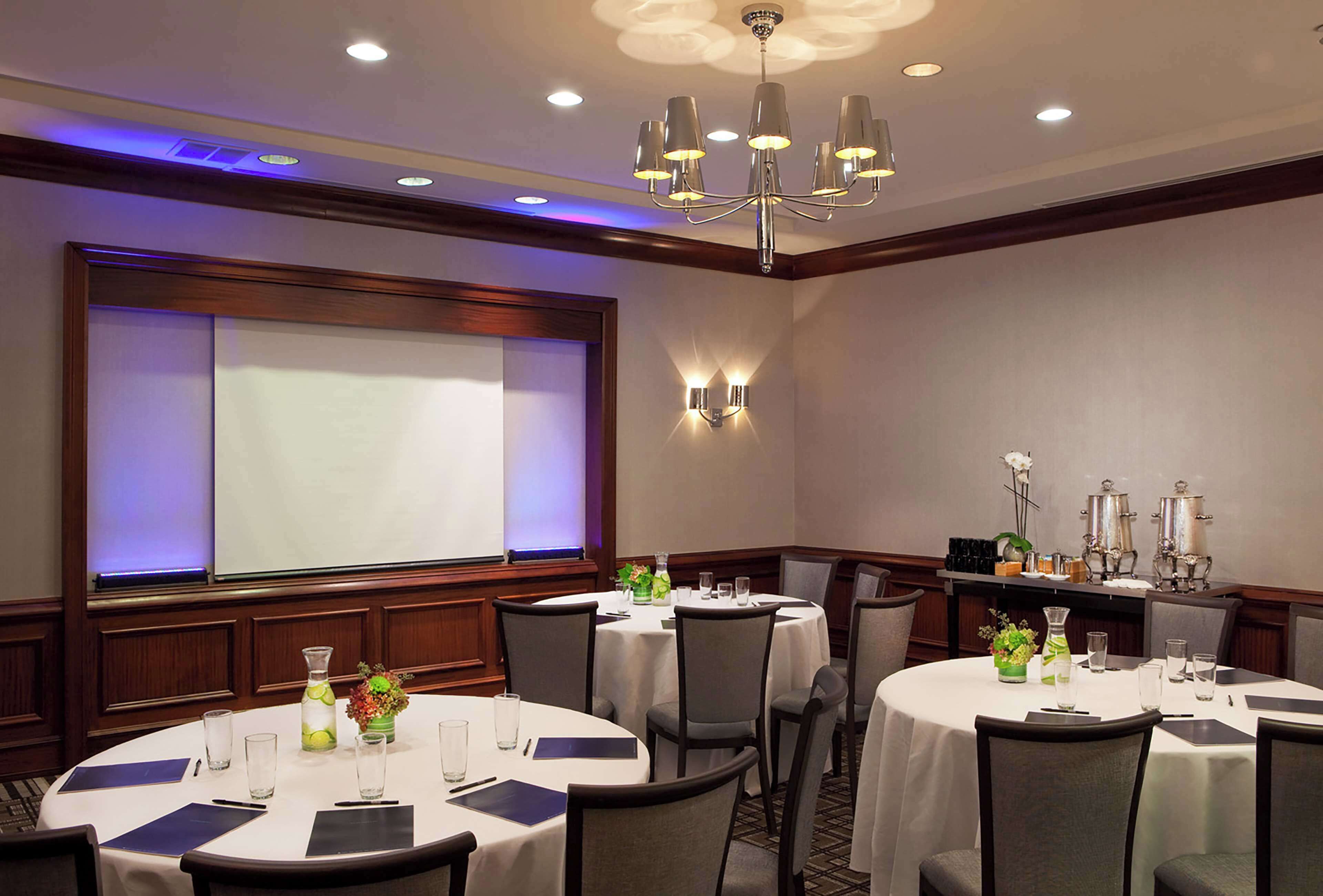 A conference room with multiple round tables, chairs, a projector screen, and a chandelier.