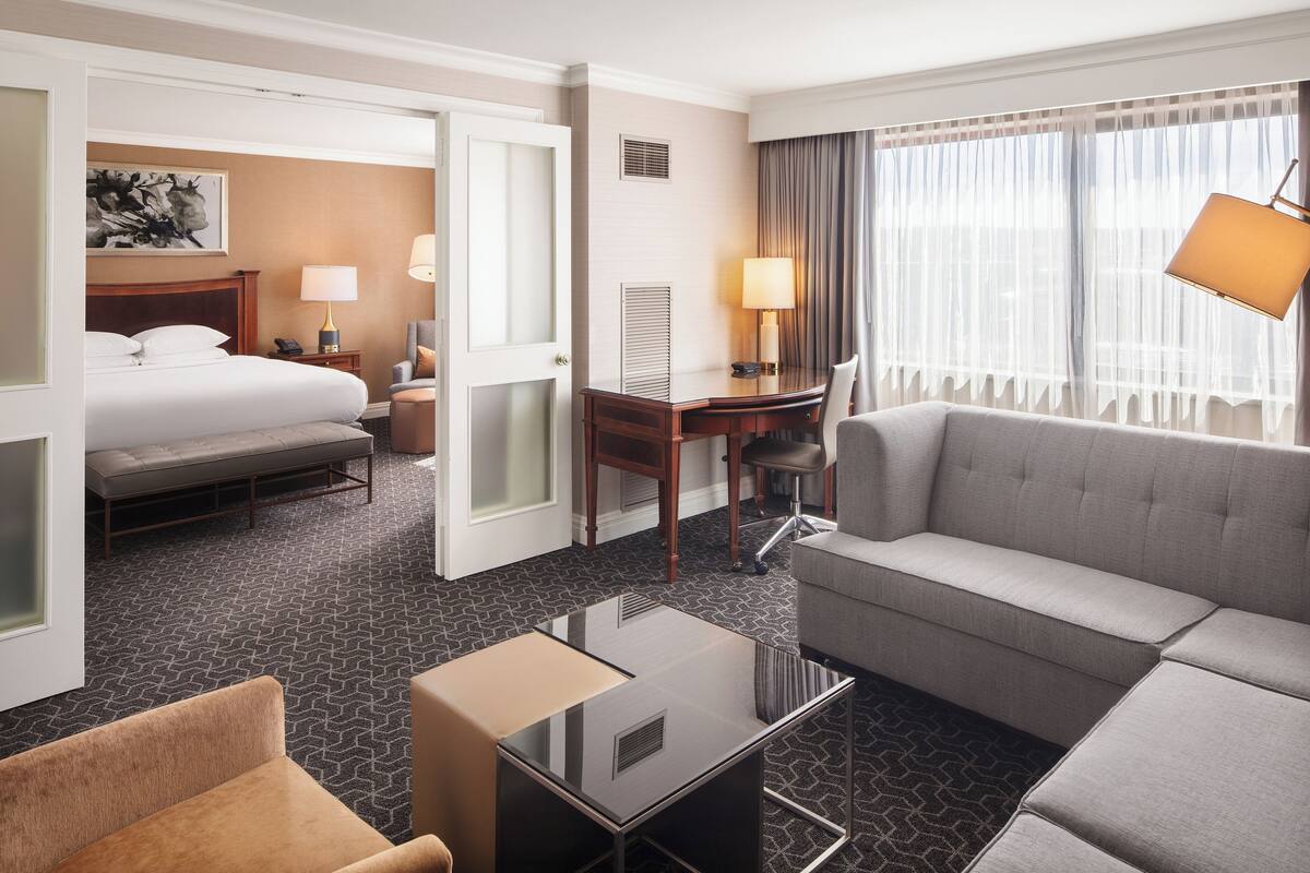 A hotel room with a bed, a sofa, a desk, and a chair. There is a window with curtains and a lamp on the desk.