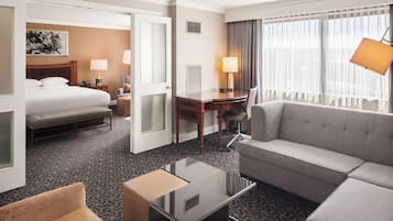 Habitación de hotel con cama, sofá, escritorio y silla. Se observa una ventana con cortinas y una lámpara en el escritorio.