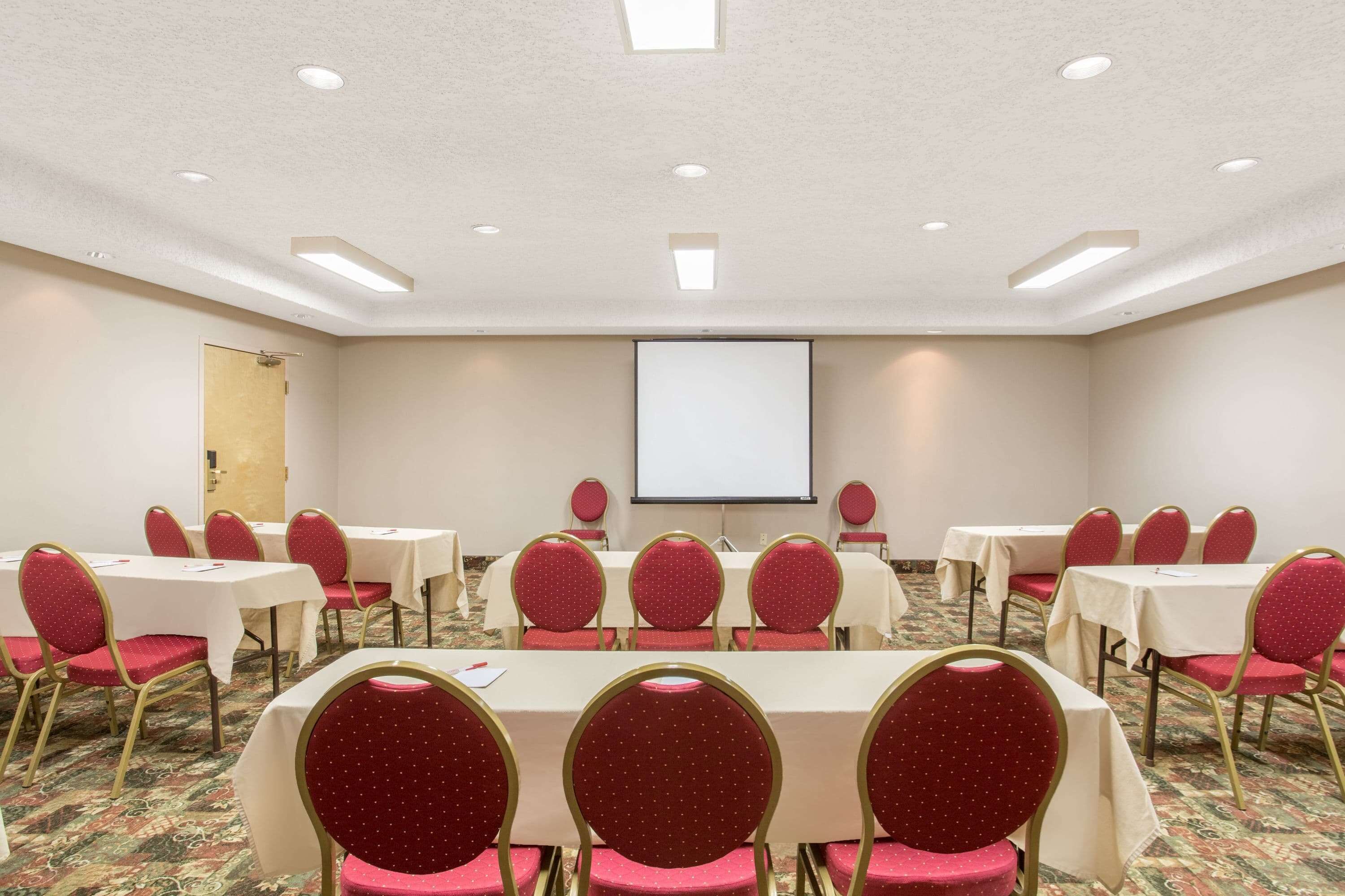 A conference room with tables and chairs, a projector screen, and a carpeted floor.