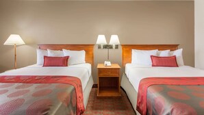 A hotel room with two beds, each with a red and grey patterned bedspread, white pillows, and a wooden headboard. There is a nightstand with two lamps between the beds.