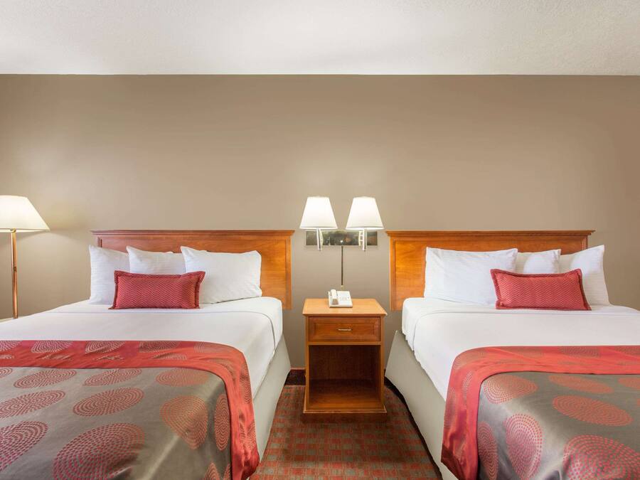 A hotel room with two beds, each with a red and grey patterned bedspread, white pillows, and a wooden headboard. There is a nightstand with two lamps between the beds.