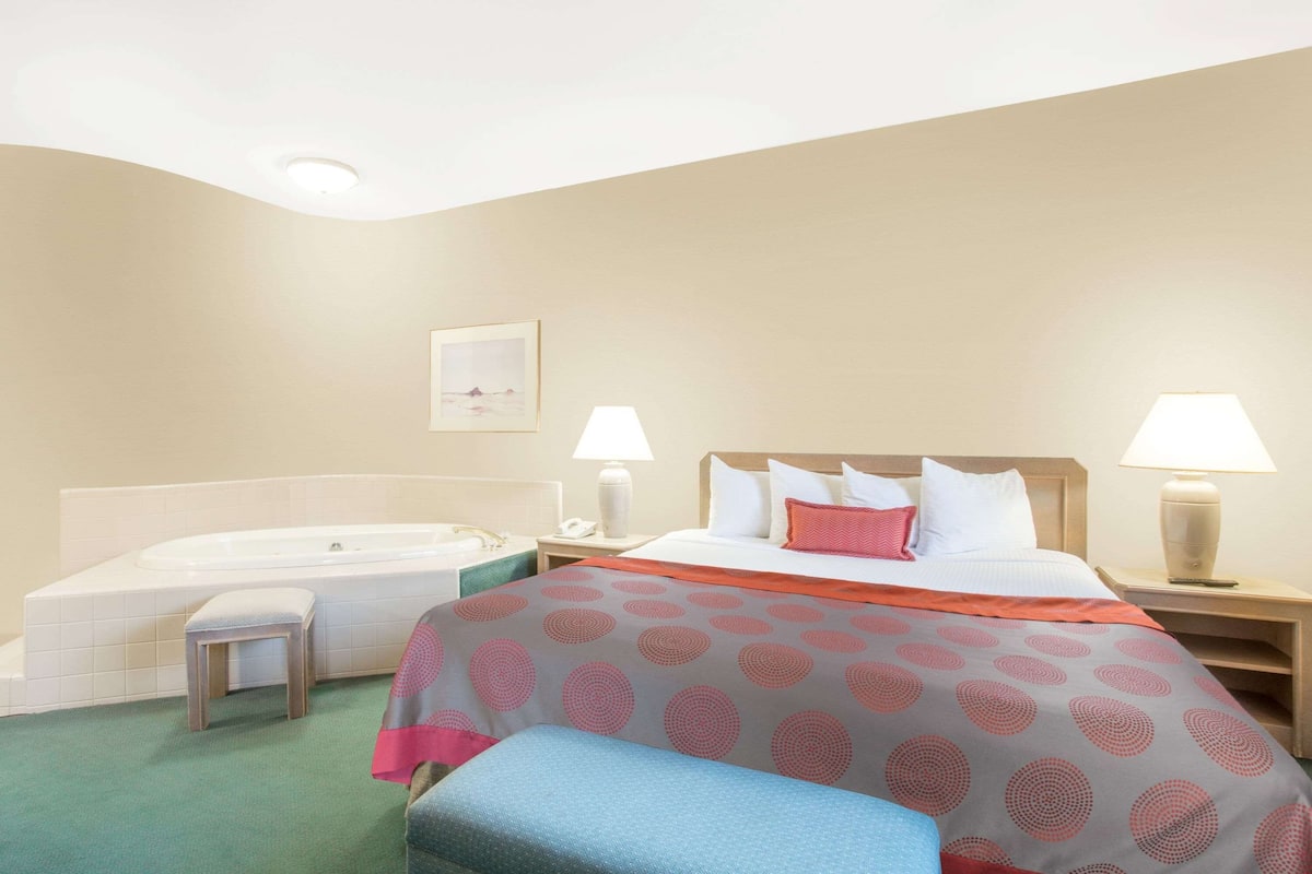 A hotel room with a bed, two bedside lamps, a nightstand, a small stool, and a bathtub.