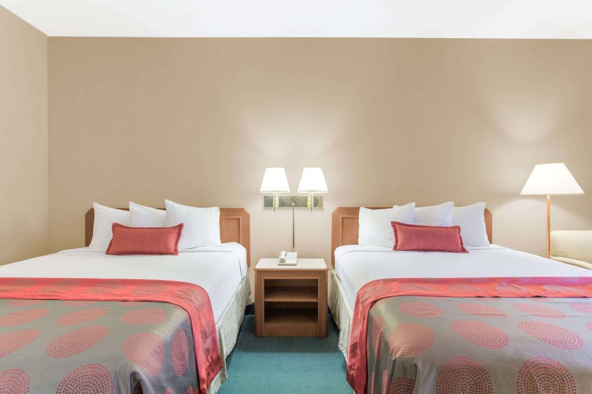 A hotel room with two beds, each with a red and grey patterned bedspread, white pillows, and a nightstand with a lamp between the beds.