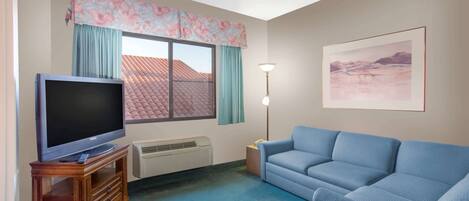 A living room with a flat-screen TV on a wooden stand, a blue sofa, a framed landscape painting, and a window with a floral valance.
