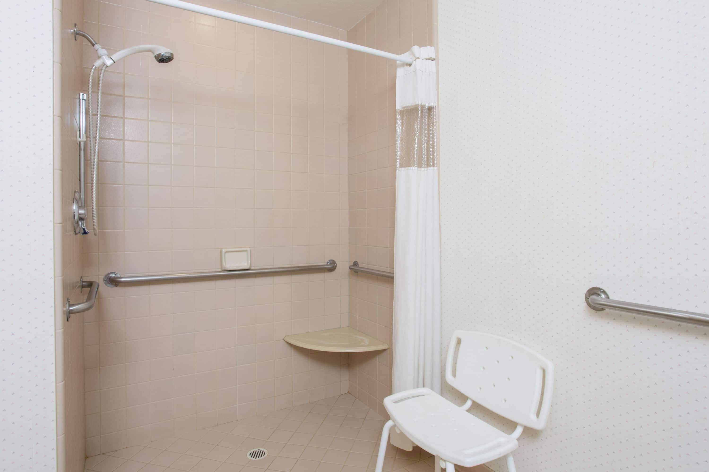 A bathroom with a shower, grab bars, a toilet, and a chair.