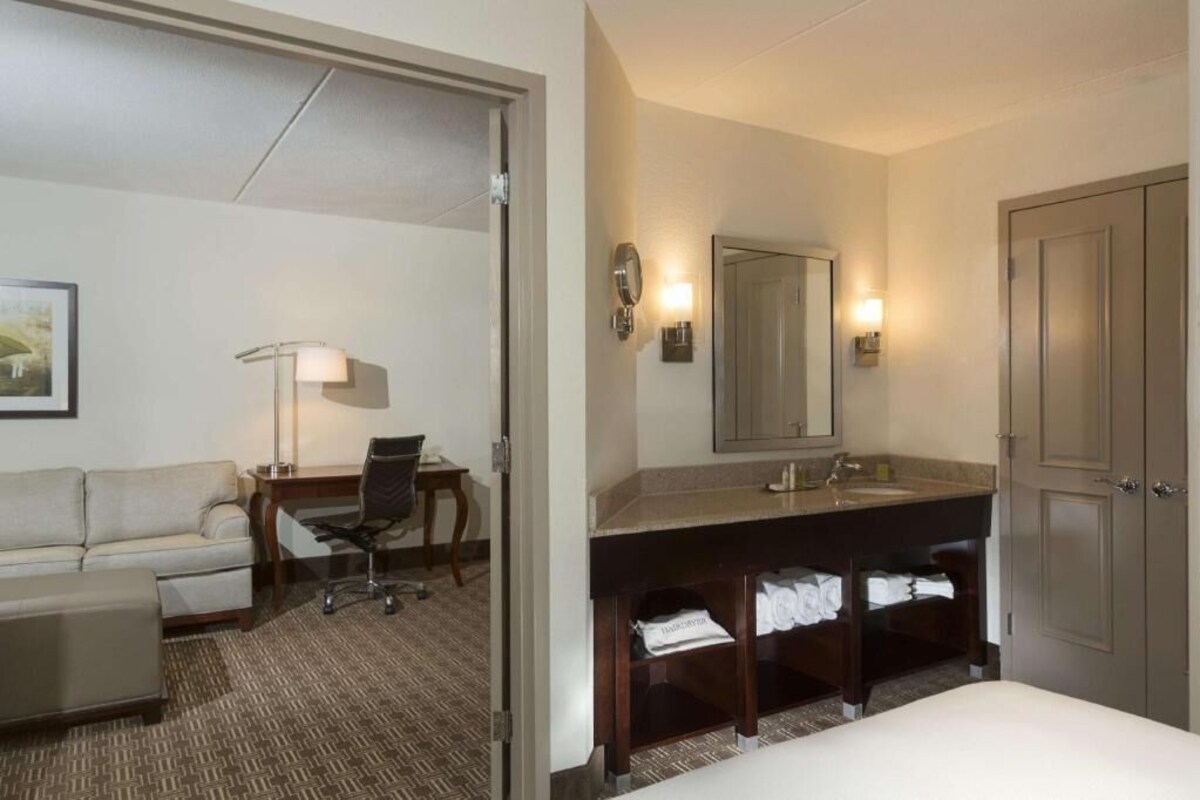 A hotel room with a bed, a desk with a chair, a sofa, a bathroom with a sink and mirror, and a door.