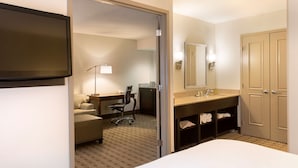 A hotel room with a bed, a desk with a chair, a desk lamp, a mirror, a sink, and a wardrobe.