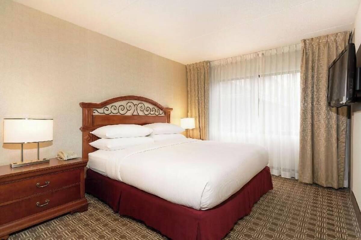 Een hotelkamer met een groot bed, een houten hoofdeinde, een nachtkastje met een lamp, een televisie aan de muur en een raam met gordijnen.