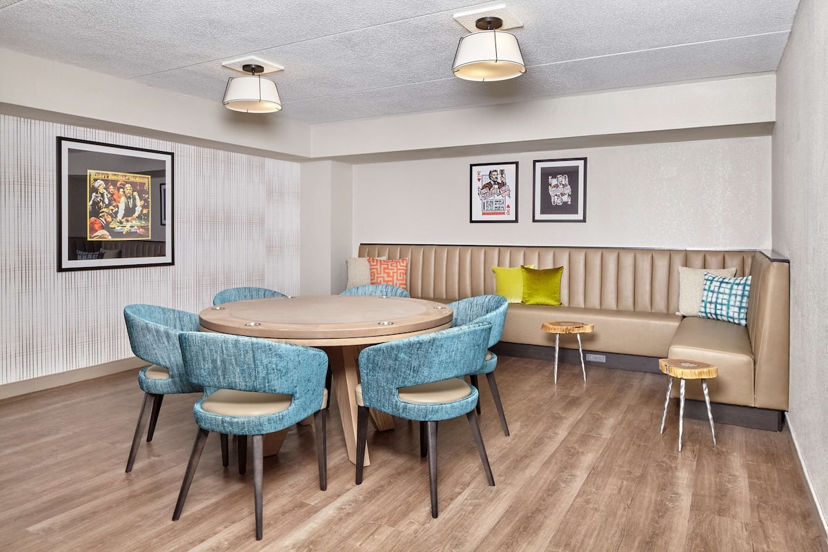 Ein modernes Zimmer mit einem runden Holztisch, blau gepolsterten Stühlen, einem beigefarbenen Sofa mit Kissen und gerahmten Kunstwerken an den Wänden.