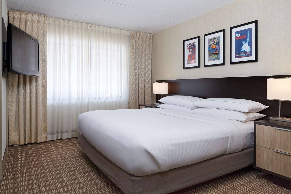 Een hotelkamer met een groot bed, twee nachtkastjes met lampen, een flatscreen televisie aan de muur en drie ingelijste posters.