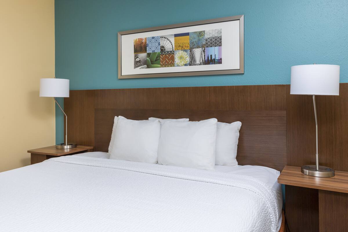Een hotelkamer met een groot bed, twee nachtkastjes, een lijst met kunstwerk boven het bed en een houten hoofdbord.