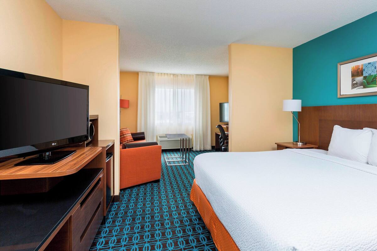 Een hotelkamer met een groot bed, een televisie op een houten standaard, een bureau met een stoel, en een bijzettafeltje met een lamp.