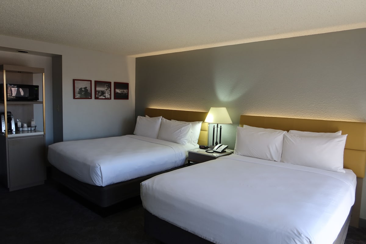 A hotel room with two single beds, a bedside table with a lamp, a small shelf with toiletries, and framed pictures on the wall.