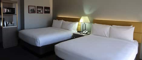 A hotel room with two single beds, a bedside table with a lamp, a small shelf with toiletries, and framed pictures on the wall.