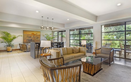 A spacious living area with a sofa, chairs, a coffee table, and a dining table. There are large windows with a view of greenery outside.