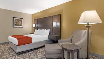 A hotel room with a bed, a chair, a table, a nightstand, and a lamp.
