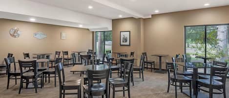 A spacious dining area with multiple tables and chairs, a large window offering a view of greenery, and wall-mounted decor including a sign and framed pictures.