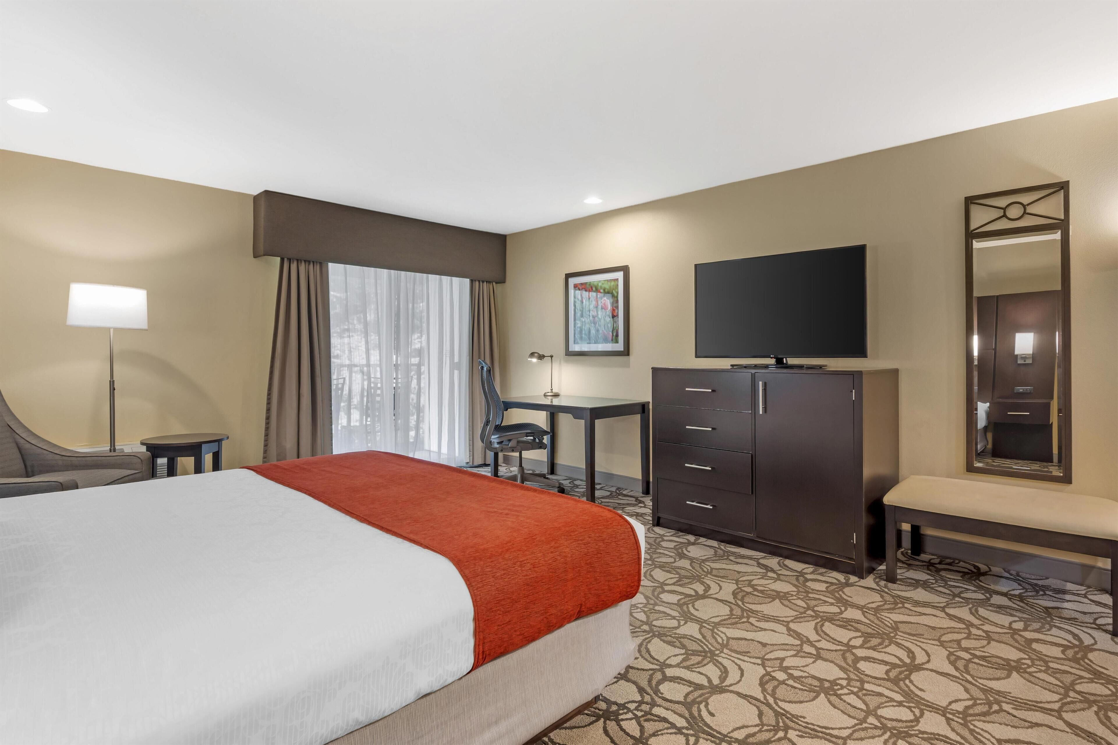 A hotel room with a large bed, a desk, a TV, a dresser, and a bench.
