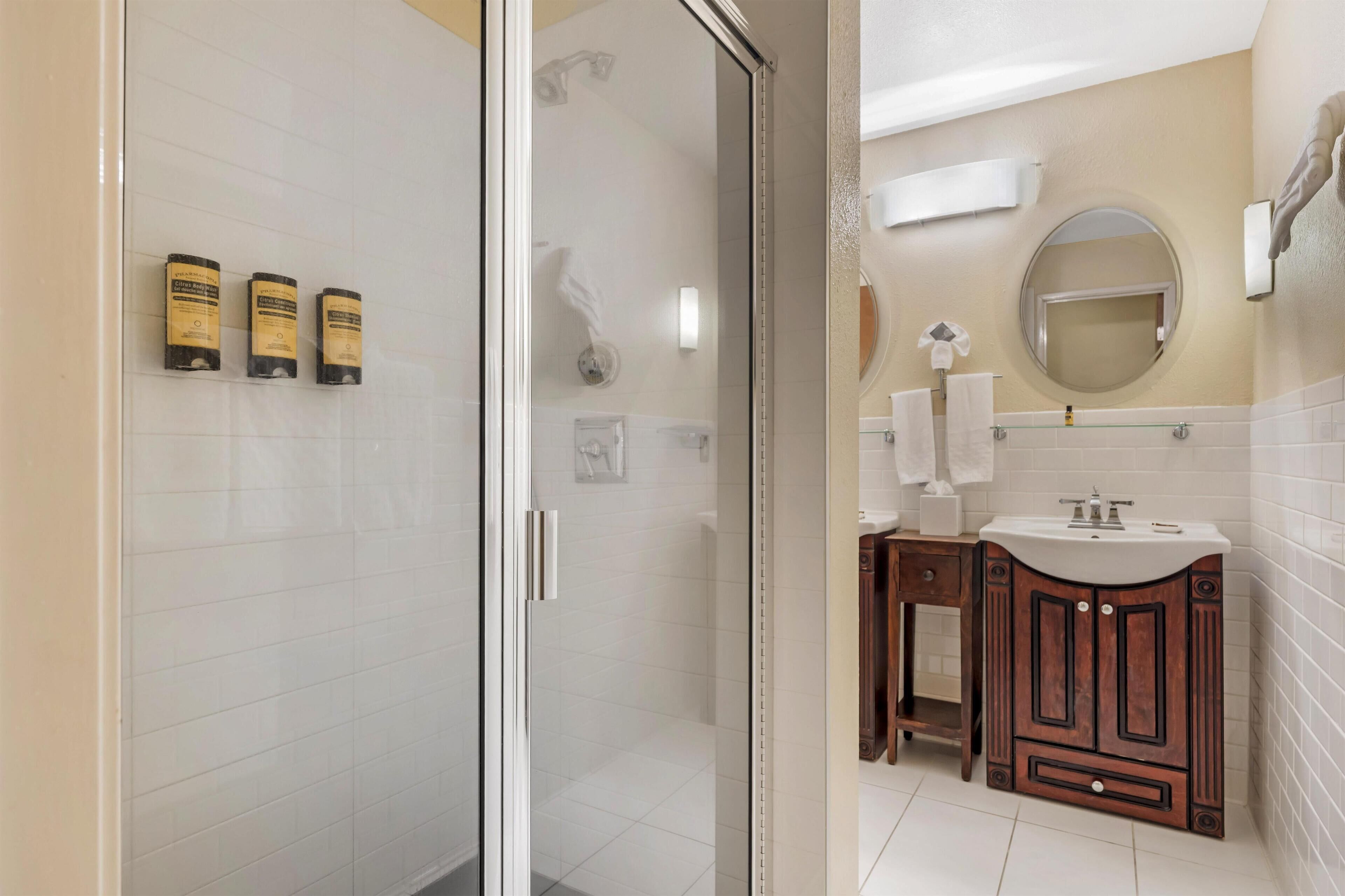 A bathroom with a glass shower enclosure, white tiled walls, a wooden vanity with a sink, and three toiletry bottles on the wall.
