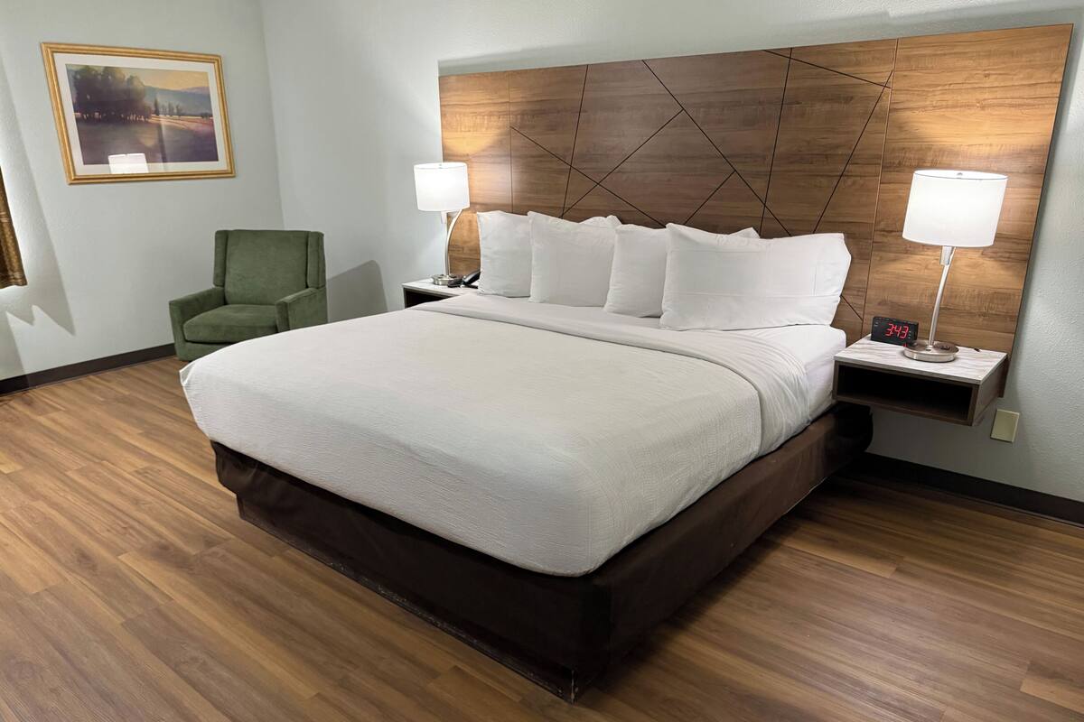 A hotel room with a large bed, two bedside lamps, a green armchair, and a wooden headboard.