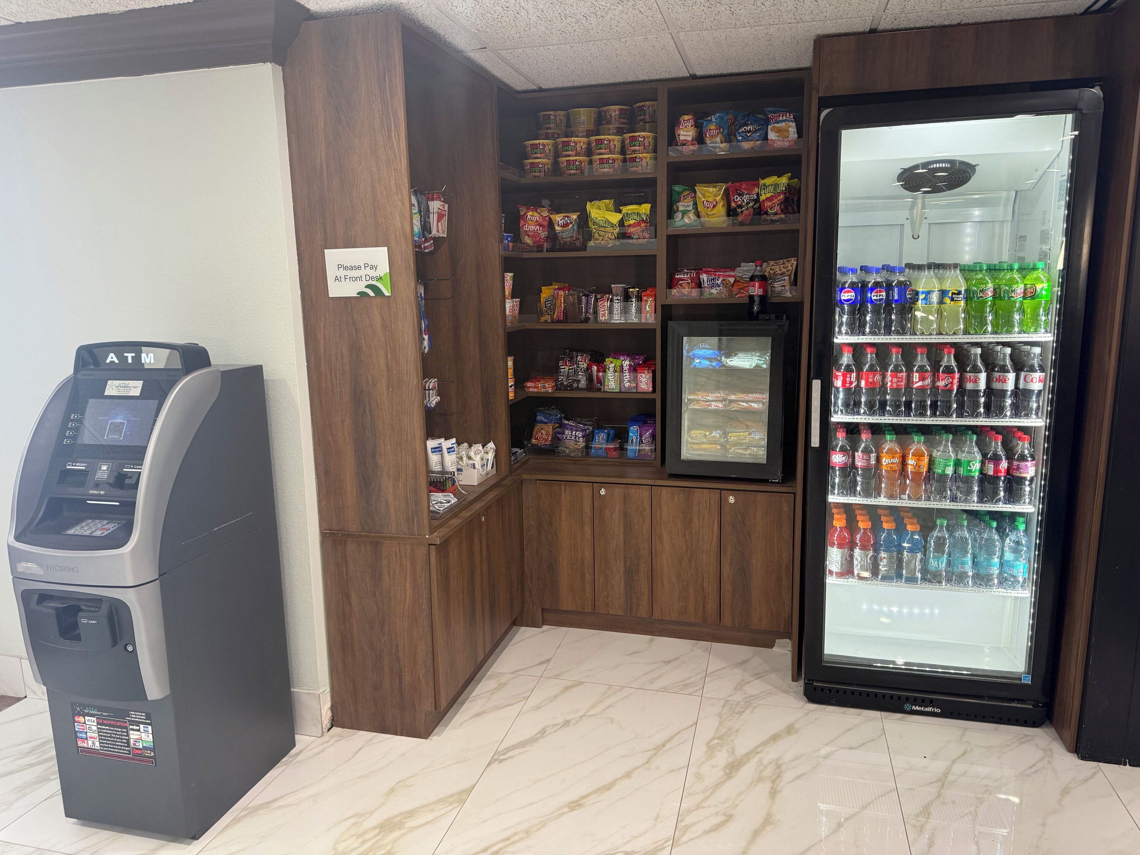 Cajero automático, estantes con aperitivos y un refrigerador para bebidas en un espacio público.