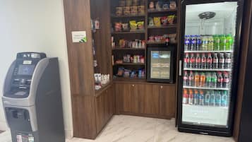 Cajero automático, estantes con aperitivos y un refrigerador para bebidas en un espacio público.