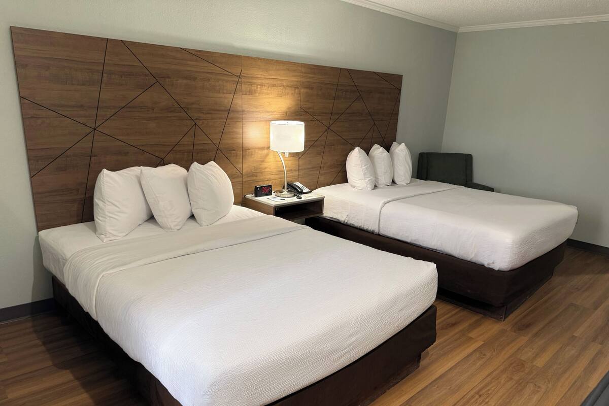 A hotel room with two beds, a wooden headboard, a bedside table with a lamp, and a digital clock.