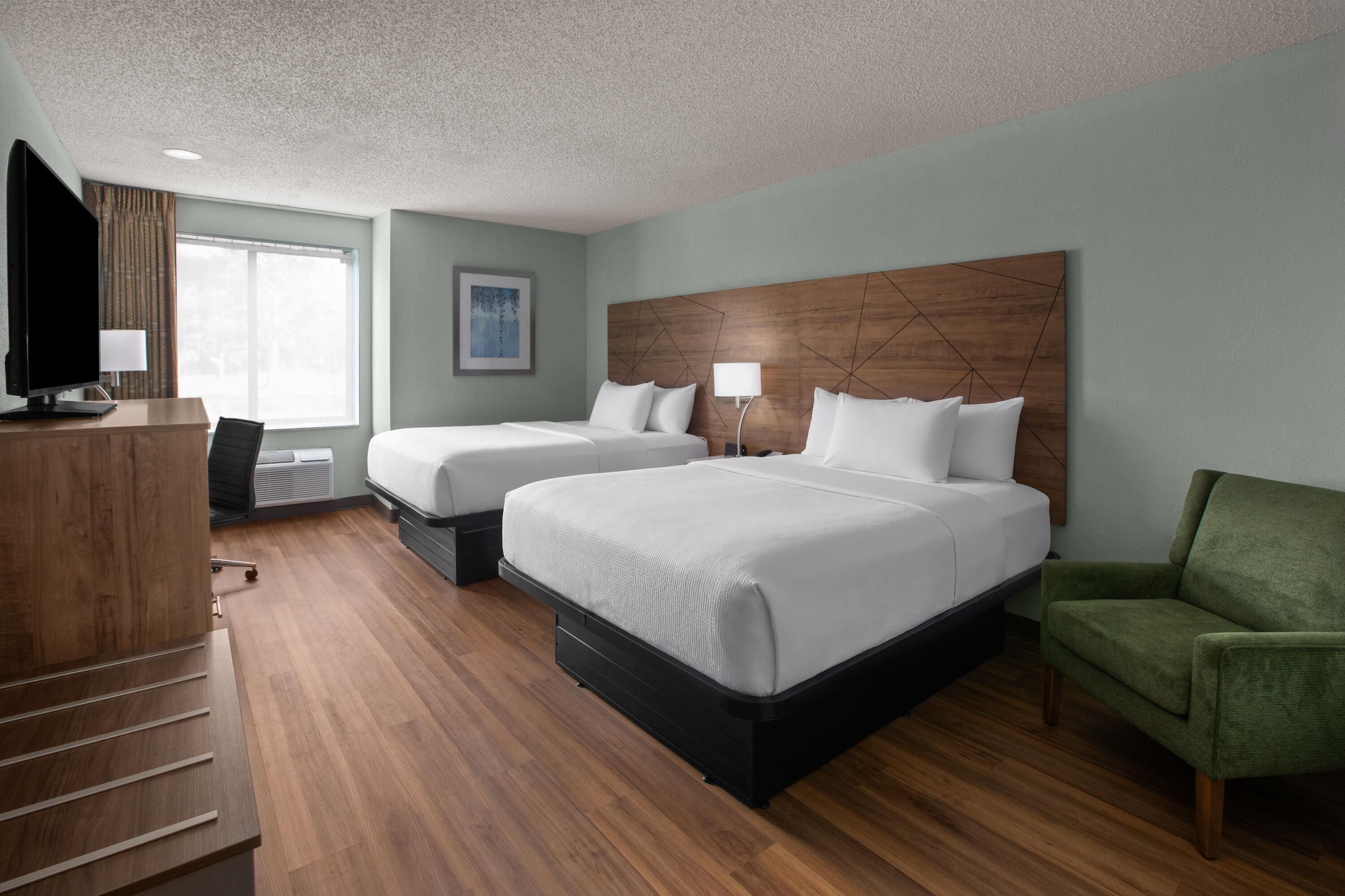 A hotel room with two beds, a desk with a flat-screen TV, a chair, and a green armchair.