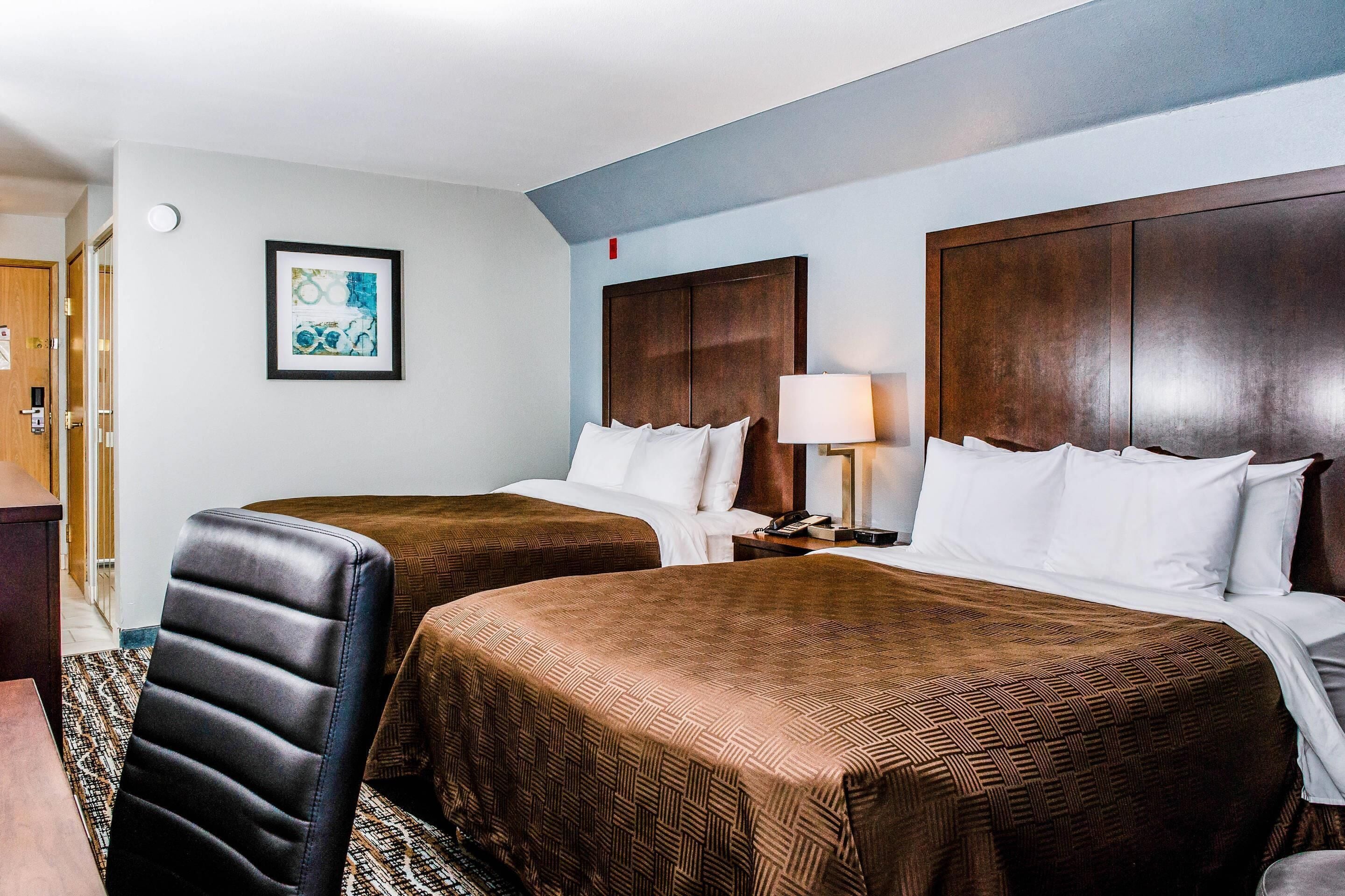 A hotel room with two beds, a wooden headboard, a framed picture on the wall, and a desk with a chair.