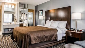 Une chambre d’hôtel avec un grand lit, une table de chevet avec une lampe, un miroir, une coiffeuse avec un lavabo et un tapis à motifs.