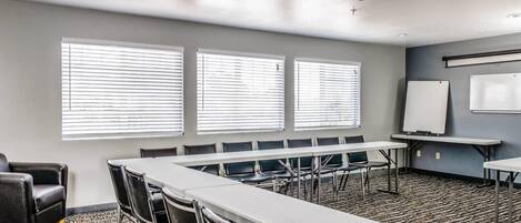 Un salón de conferencias con una mesa larga, sillas, una pizarra y ventanas amplias con persianas.
