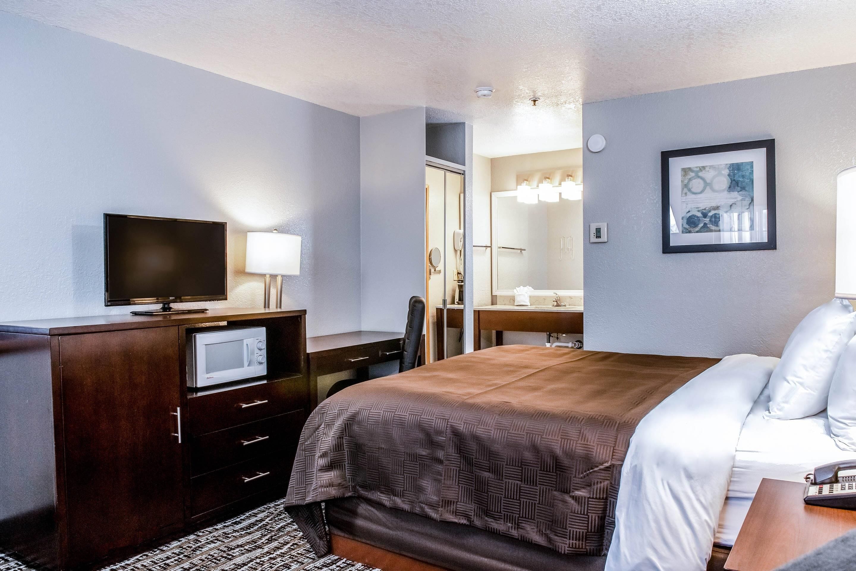 A hotel room with a bed, a desk with a computer, a television, a microwave, a bathroom with a mirror and lights, and a framed picture on the wall.