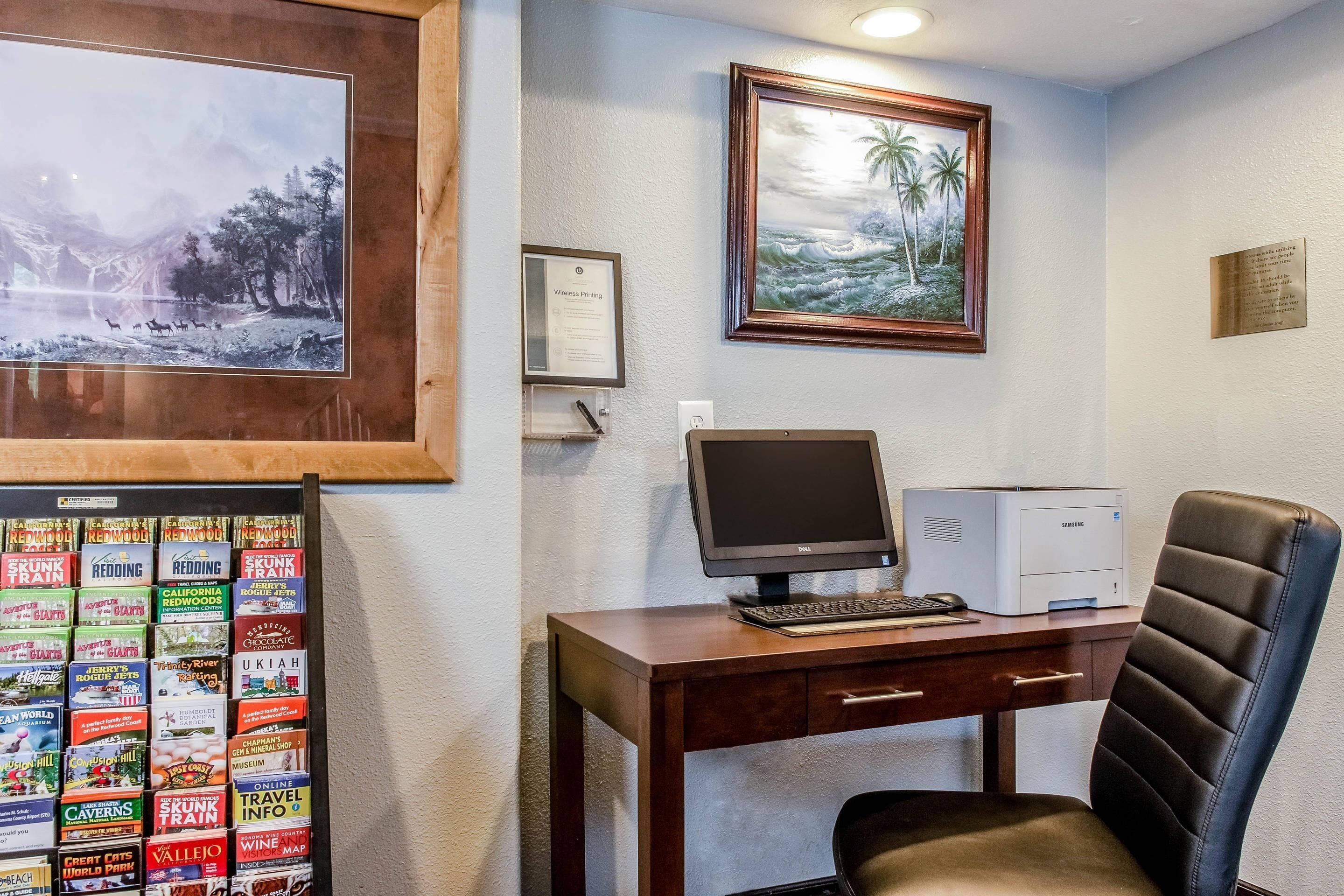 A home office with a desk, computer, chair, framed artwork, and travel brochures.