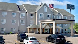 A Country Inn & Suites hotel with a two-story building, a flagpole with an American flag, and a parking lot with several cars. - Country Inn & Suites by Radisson, Owatonna, MN (Owatonna)