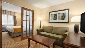 A hotel room with a bed, a sofa, a table, a lamp, and a television. - Country Inn & Suites by Radisson, Owatonna, MN (Owatonna)