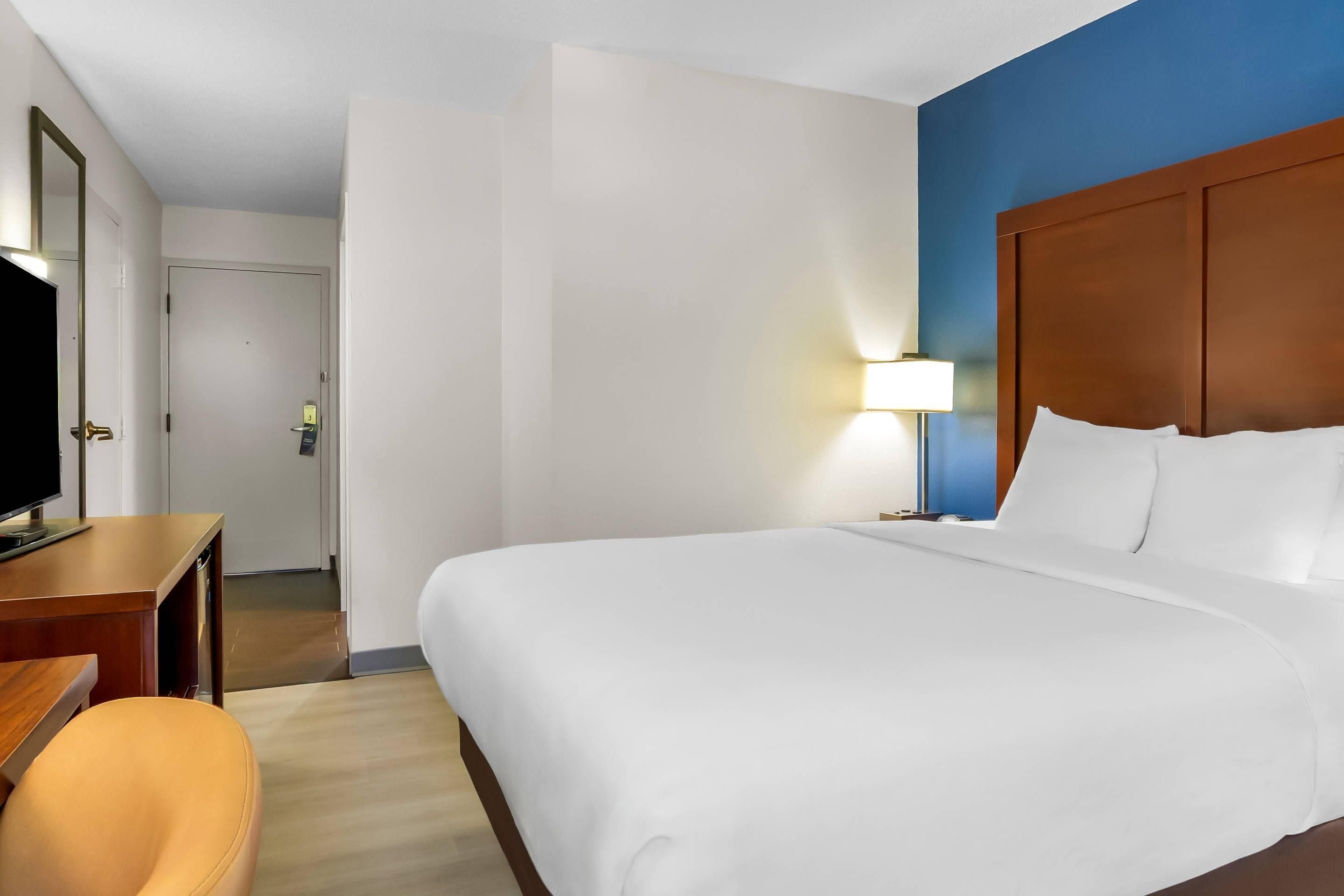 Una habitación de hotel con una cama grande, un escritorio con televisor, una silla y una lámpara.