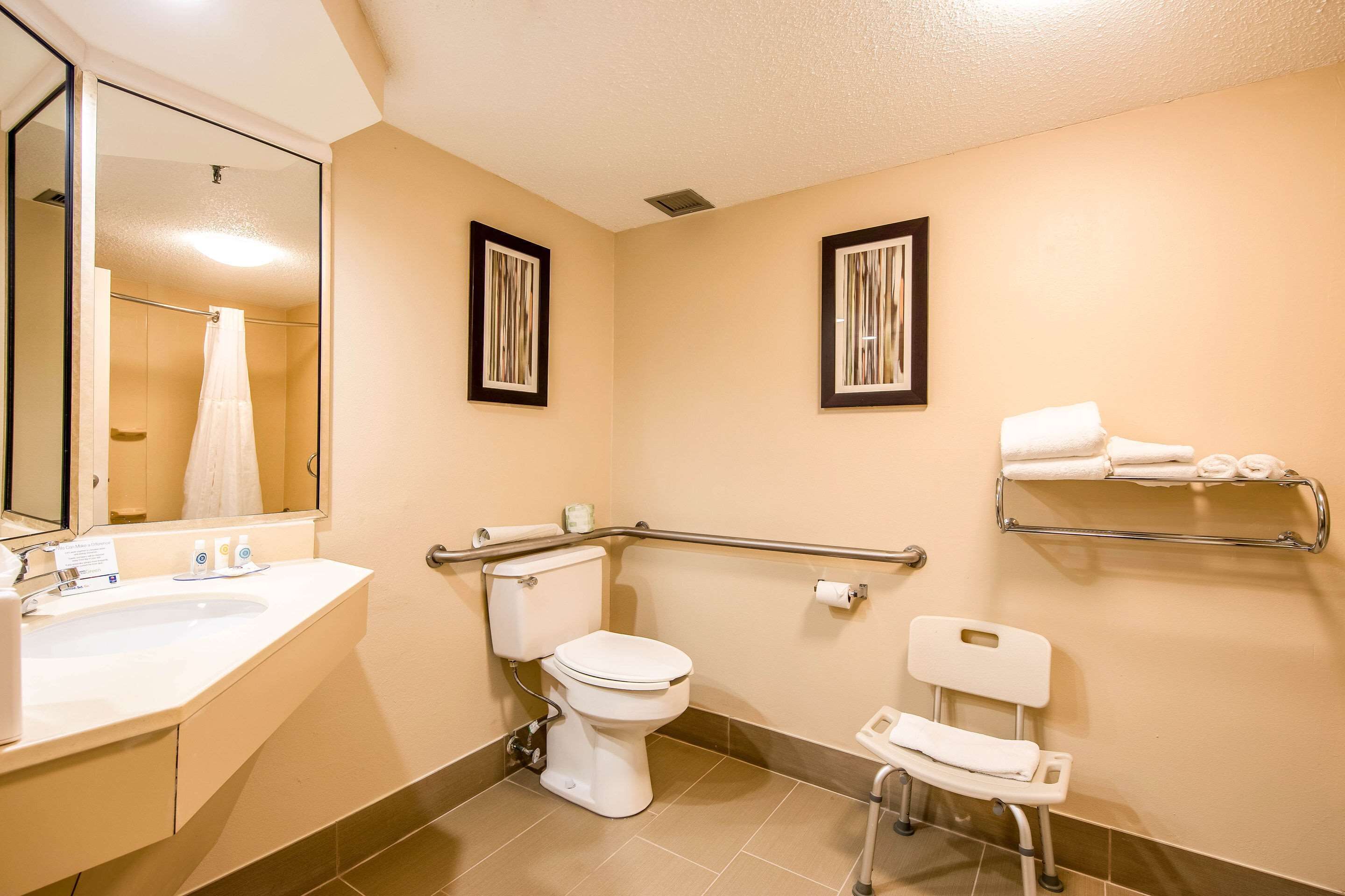 Un baño con lavamanos, inodoro y bidé blancos, un banco accesible para sillas de ruedas y un perchero con toallas.