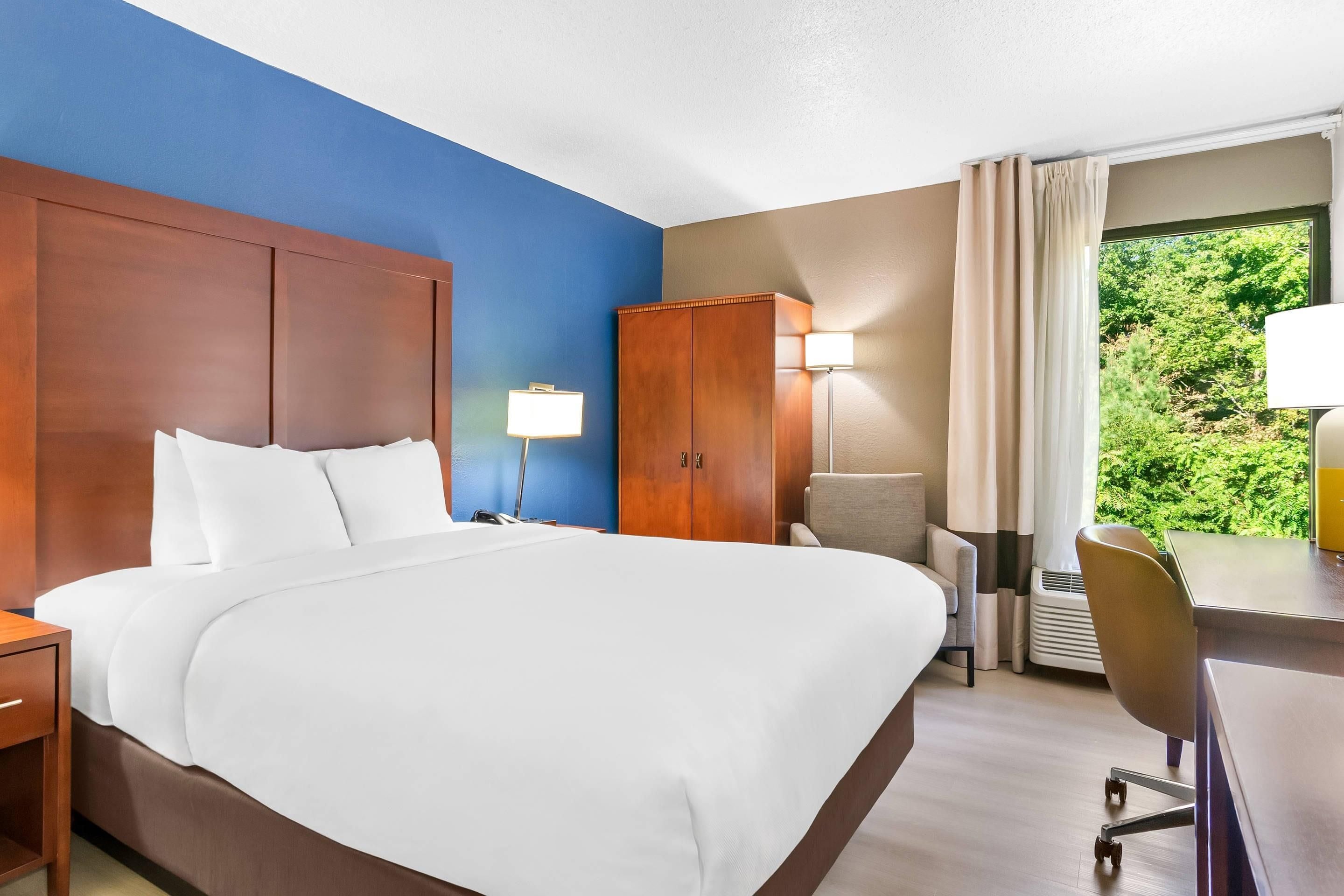 Habitación de hotel con una cama grande, un armario de madera, un escritorio con silla y una ventana con vista a zonas verdes.