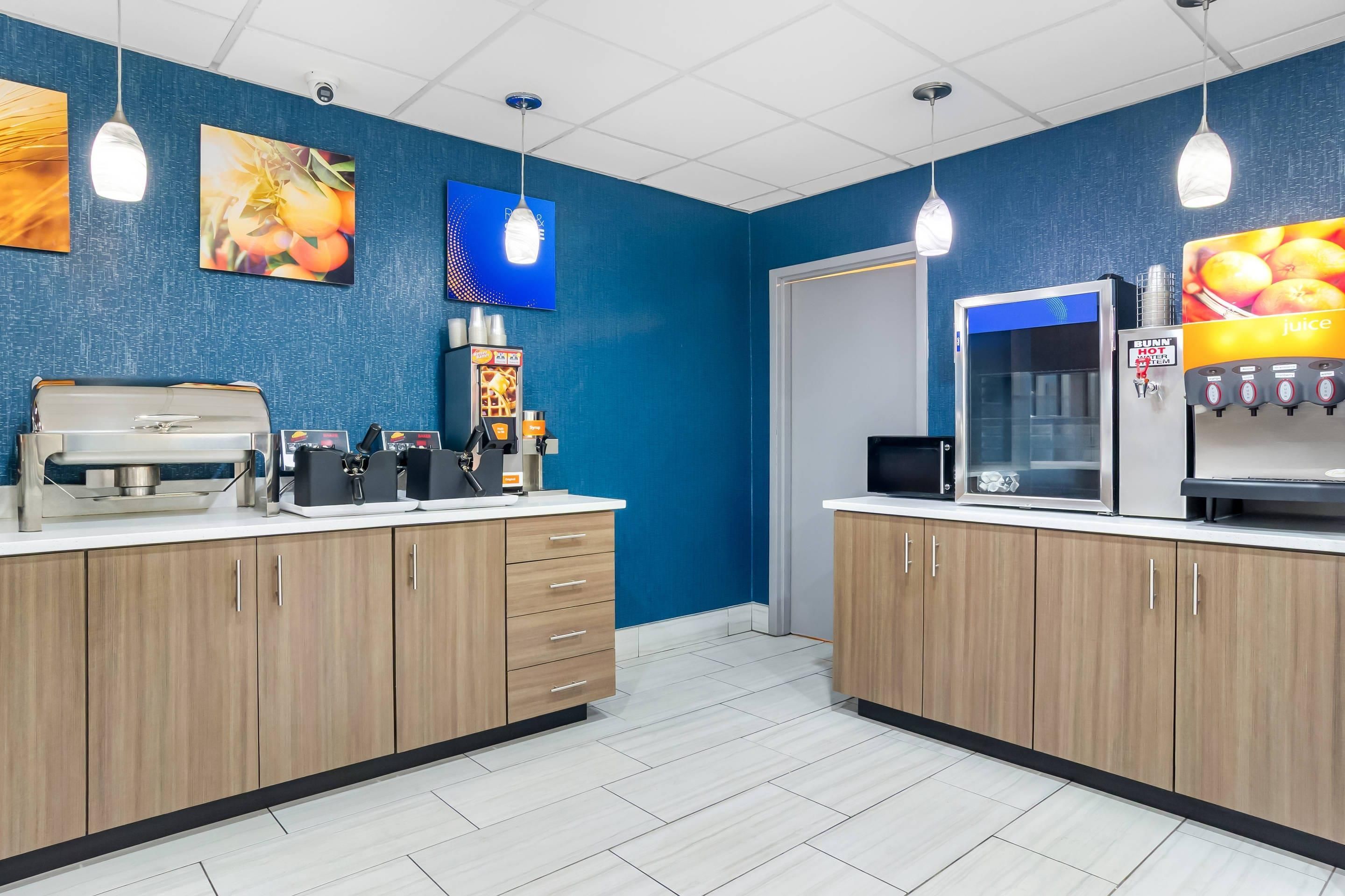 A modern kitchen area with wooden cabinets, a coffee machine, a juice dispenser, and a refrigerator.