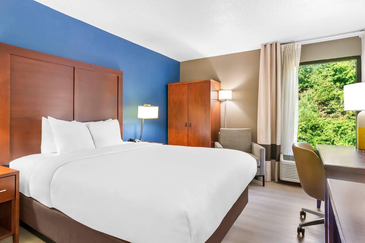 A hotel room with a large bed, a wooden wardrobe, a desk with a chair, and a window with a view of greenery.