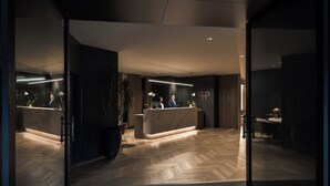 Un área de recepción moderna con piso de madera en patrón de espiga, una mesa de recepción y una zona de estar con una lámpara.