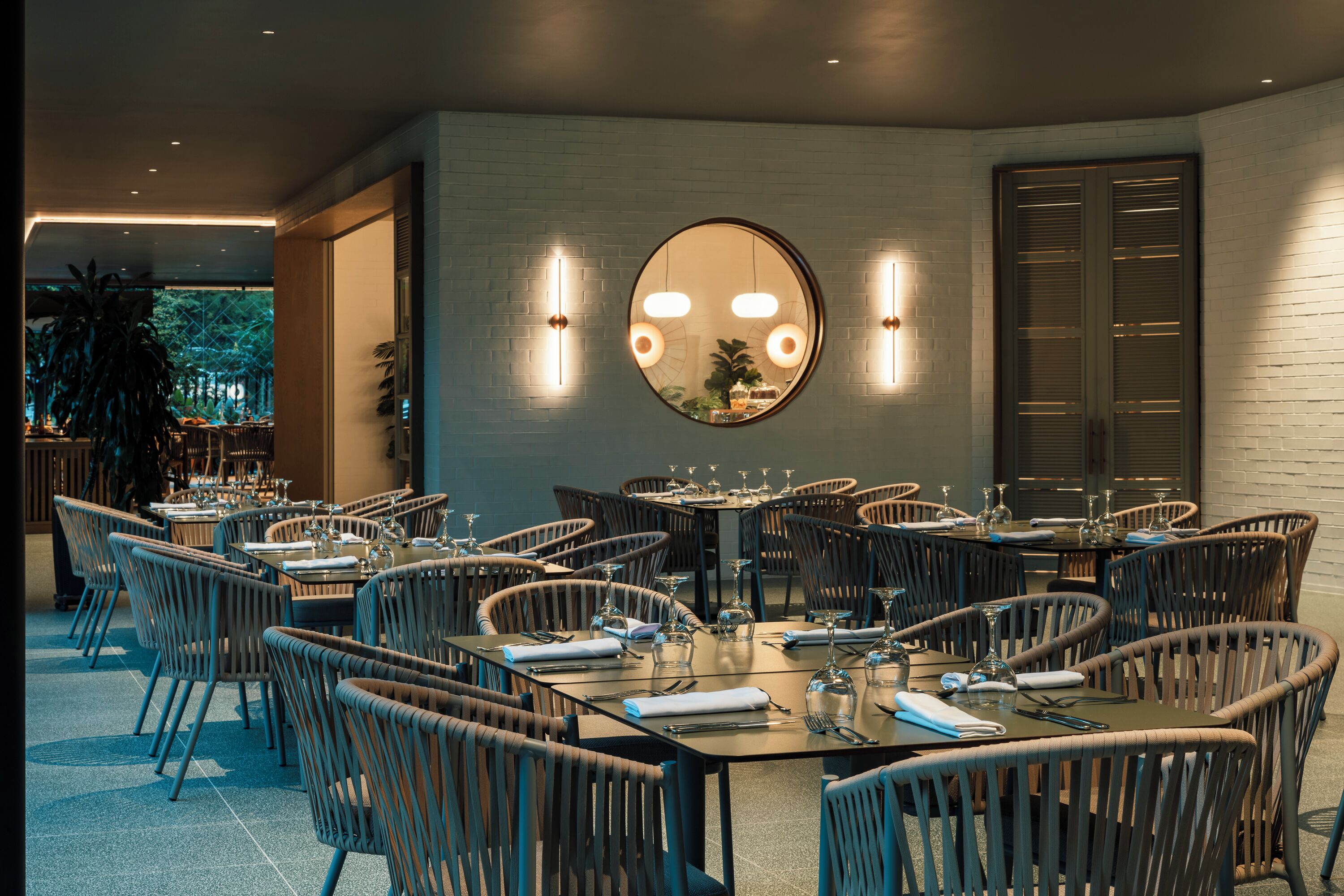 A restaurant with round tables and wicker chairs, a large mirror, and wall-mounted lights.
