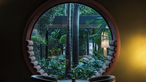 Ventana circular con vistas a un jardín exuberante, una mesa redonda y dos sillas.