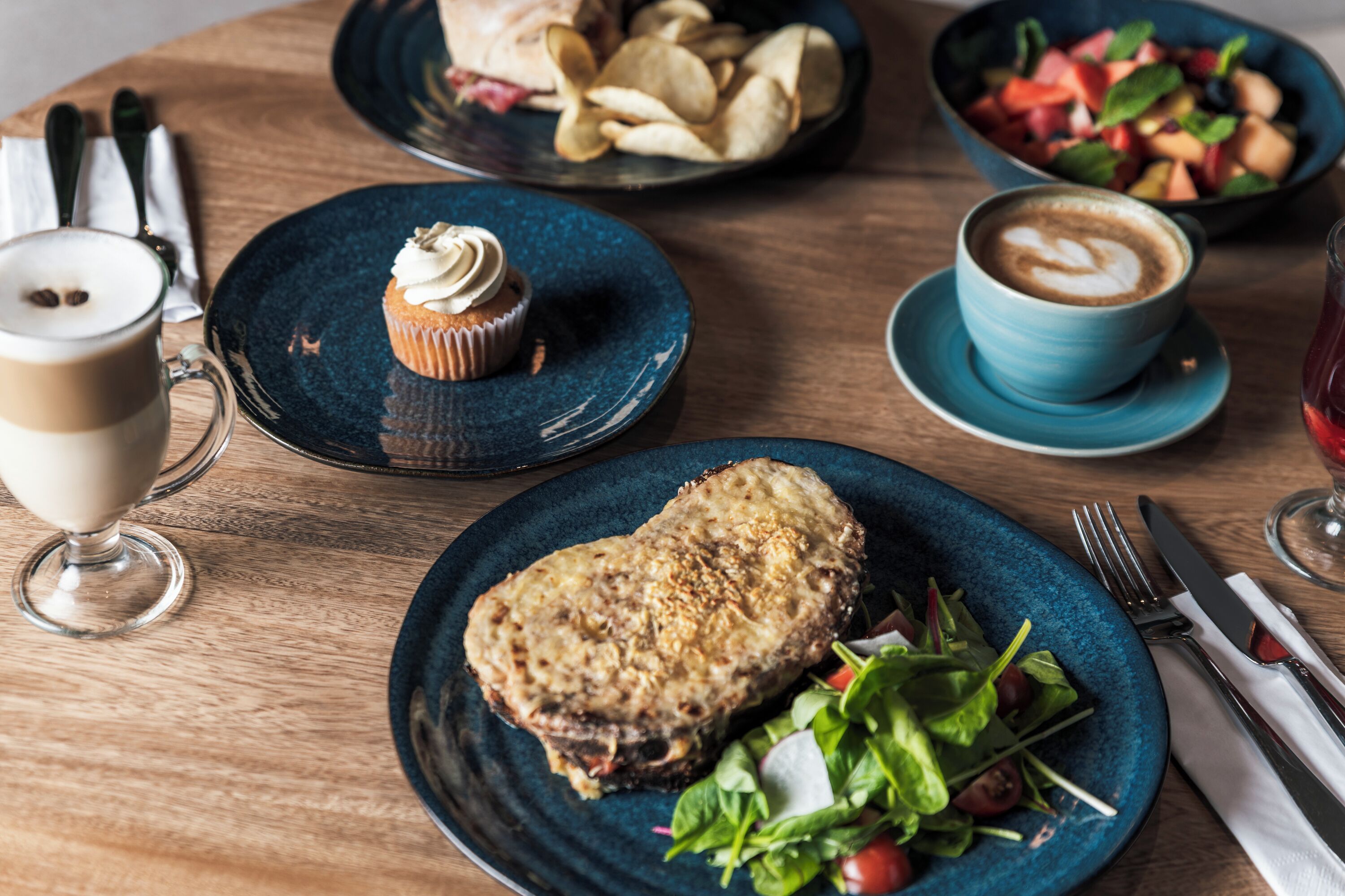 A wooden table with a variety of food items including a plate with a sandwich, a bowl of salad, a plate with a cupcake, a cup of coffee, and a glass of red wine.