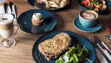 A wooden table with a variety of food items including a plate with a sandwich, a bowl of salad, a plate with a cupcake, a cup of coffee, and a glass of red wine.