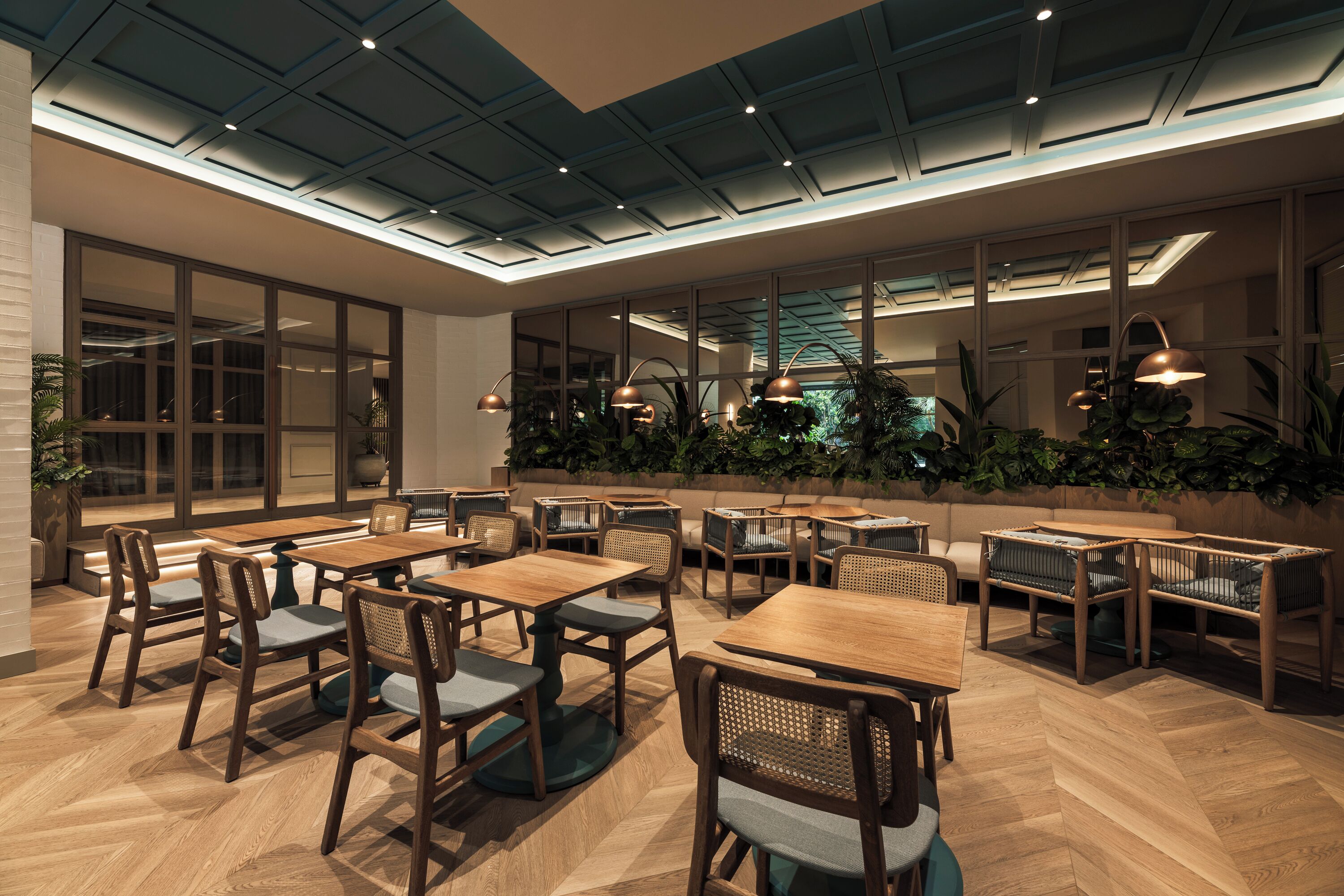 Un restaurant avec des tables et des chaises en bois, un plafond haut doté d’un éclairage encastré et de grandes fenêtres ornées de plantes.
